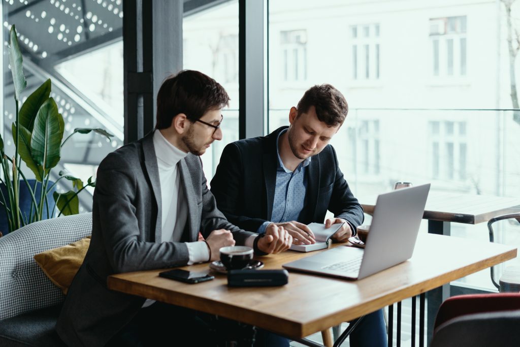 Two businessmen talking about new opportunities sitting with laptop at desk near large window, planning project, considering business offer, sharing ideas while drinking coffee together